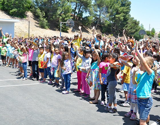 A line of children in a schoolyard raising their hands.